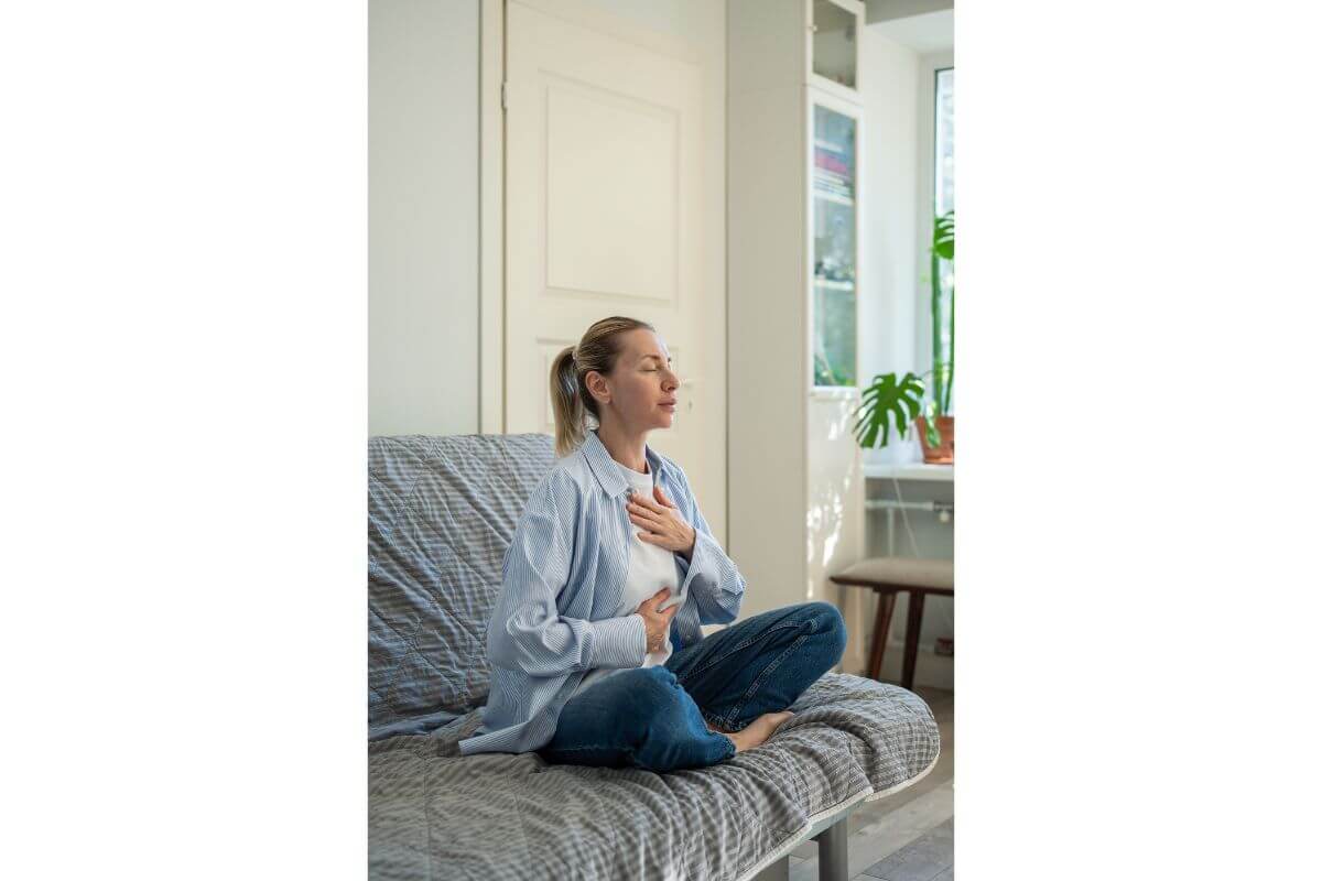 Frau sitzt ruhig auf dem Sofa und legt die Hände auf Brust und Bauch – Atemübungen helfen, das Nervensystem zu regulieren.