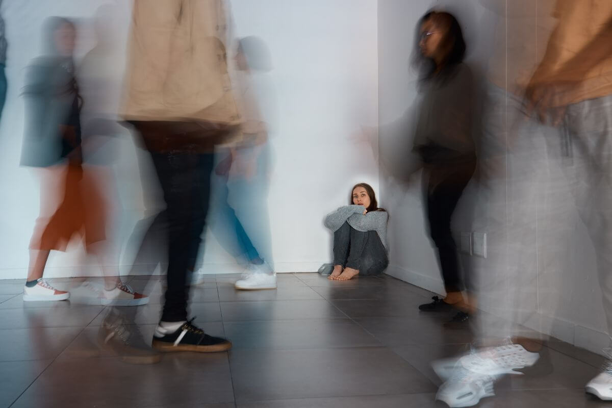 Frau sitzt zurückgezogen an einer Wand, während Menschen vorbeigehen – Sinnbild für Rückzug und Überforderung bei sozialer Phobie