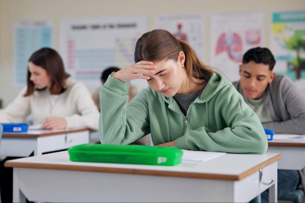 Junge Frau sitzt in einer Prüfungssituation und wirkt angespannt, hält die Hand an die Stirn. Sie versucht sich zu konzentrieren, während andere Personen im Hintergrund arbeiten.